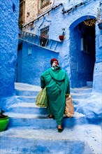 An Elderly Woman Walks Through The Colourful Streets Of The Medina With Heavy Shopping Bags, Chefchaouen, Morocco