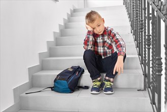 Front view of sad blond boy sitting on stairs in corridor of school and leaning head by hand. Little disappointed pupil thinking about his bed behavior on lesson. Concept of study.