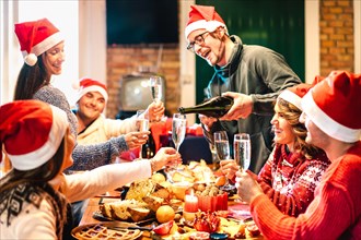 Millenial friends on santa hats celebrating Christmas with champagne and sweets food at log cabin - Winter holidays concept with young people