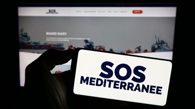 Person holding cellphone with logo of French sea rescue organisation SOS Mediterranee in front of webpage. Focus on phone display.