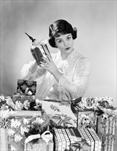 1950s WOMAN WEARING NIGHTGOWN CHRISTMAS MORNING SITTING BY PILE OF CHRISTMAS PRESENTS HOLDING SHAKING ONE PRESENT
