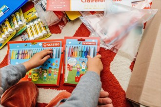 Paris, France - Sep 1, 2023: From the viewpoint of a toddler, we see small hands holding packages of Stabilo colored pencils during the unboxing of back-to-school items