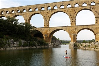 Woman in bikini and shorts stand up paddling (SUP) on the Gardon river in front of Pont du Gard, Provence, France.