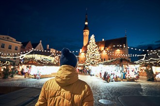 Rear view of man in warm clothing during night walk in city. Traditional Christmas market on Town Hall Square in Tallinn, Estonia.