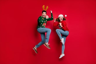 Full body photo of amazed jumping couple excited by x-mas prices wear ugly ornament jumpers and headwear isolated red color background