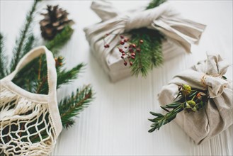 Stylish christmas gifts wrapped in linen fabric with green branch and reusable shopping bag with green spruce on rustic wooden background. Zero waste