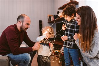 happy family christmas time at home. smiling mother and father with cute children decorating christmas lights together. family unit and christmas spir