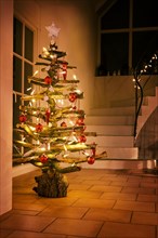 Rustic Christmas tree made of raw wood branches decorated with glowing fairy lights and red balls in a stairwell at night, selected focus, narrow dept