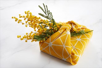 Wrapped gift by linen fabric in furoshiki eco style with yellow mimosa flower. Woman or mother day celebration concept.