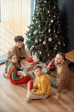 Portrait of positive beautiful Caucasian family sitting with presents on floor against Christmas tree at home