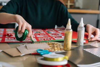 Close-up hands of unrecognizable young woman using holiday ribbon to frame board with advent calendar envelopes at home.