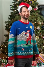 Portrait of a young man with a beard, wearing a Santa Hat and an ugly Christmas sweater standing in front of Christmas tree.