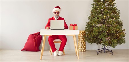 Funny modern Santa working on laptop while sitting at desk in office with Christmas tree