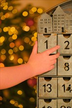 Metallic advent calendar and childs hand near Christmas tree with glowing garland on blurred room background.Christmas traditions and symbols. Gifts