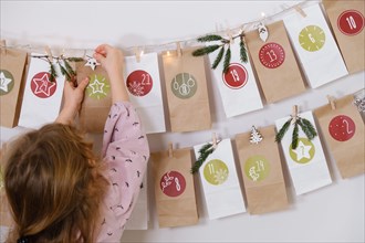 Toddler child gets new advent calendar task, waiting for Christmas. Festive mood for cute curly girl. Kid takes advent bag with dates from the wall at