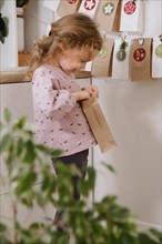 Toddler child gets new advent calendar task, waiting for Christmas. Festive mood for cute curly girl. Kid takes advent bag with dates from the wall at