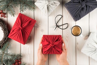 Female hands hold christmas present in furoshiki package, top view. Spruce branches, scissors, cup of coffee and gift boxes on planking. Christmas or