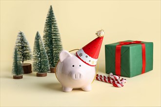 Piggy bank in festive party hat with Christmas trees, gift box and candy canes on beige background