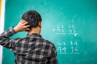 Confused kid scratching head to solve mathematics problem on board at classroom - concept of intelligence, trouble learning and education