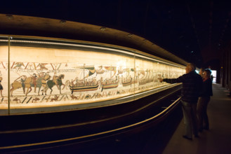 A couple of tourists admiring the Bayeux tapestry or La Tapisserie de la Reine Mathilde in it's current display case