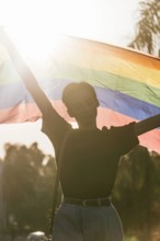 Delighted male gay standing with eyes closed raising rainbow LGBT flag during sunset in city