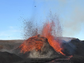 Volcanic vent at Fagradalsfjall, Iceland, erupting incandescent orange and red lava. Black lava in the foreground and blue sky in the background.