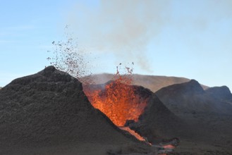 Volcanic vent at Fagradalsfjall, Iceland, erupting incandescent orange and red lava. Black cool lava in the foreground and blue sky in the background.