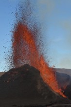 Volcanic vent at Fagradalsfjall, Iceland, erupting orange and red lava. Black cooled lava in the foreground and blue sky in the background.