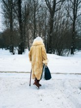 Winter. Back view of elderly women in fur coat with bags walking along snow with cane. Seniors, lifestyle concept