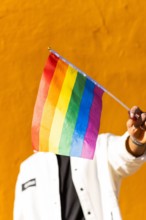 A young adult holds a multicolored rainbow flag in front of a bright yellow background