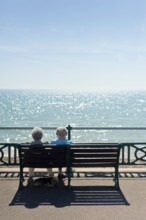 Elderly old couple on a bench enjoying the sun at the seaside while their dog lies underneath in the shade, UK