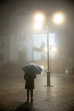 Person with umbrella standing under street lamp in front of the duomo in rain and fog at night, Ravello, The Amalfi Coast, Campania, Italy, Europe