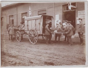 Wounded soldier on a stretcher, being loaded by stretcher bearers into a horse drawn ambulance. They are parked outside a building with Red Cross flags, the road is a wide mud track.