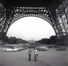 1961, historical, cars of the era parked underneath the Eiffel Tower in Paris, France, with two men standing in the open area under the giant metal structure, built for the 1889 Paris Exposition. Orig...