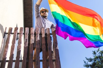 Man in hat holding lgbt rainbow flag in his hands while standing on the balcony.