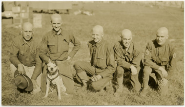 Horizontal, sepia photograph showing five uniformed soldiers kneeling in a grass field posing for a picture. One of the soldiers is holding the leash of a dog wearing a harness and a collar. Title: Fi...