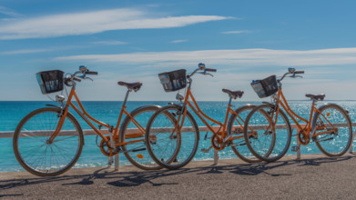 Three bright orange hire bicycles parked on the seaside promenade with an azur blue sea sparkling in the background