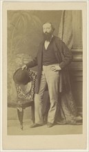 bearded man standing with one hand on hip, the other with hat in hand on chair back; F. Schwarzschild, British, active Calcutta