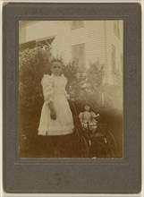 little girl in white dress standing next to a baby carriage with a doll inside; American; about 1900; Gelatin silver print