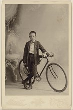 young boy with cap in hand, standing with bicycle, in a studio setting; Hewitt, American, active Nunda, New York 1880s - 1890s