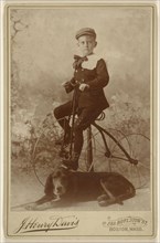 little boy wearing a cap, mounted on an old-style three-wheel bicycle, with a dog in the foreground; J. Henry Davis, American