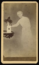 Female  spirit  standing next to a table with a photograph propped against a vase with flowers; William H. Mumler, American