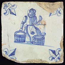 Figure tile, blue with seated saleswoman with two barrels of goods and scale in the hand, corner motif lily, wall tile