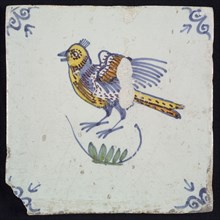 Animal tile, bird above branch to the left in yellow, brown, green and blue on white, corner pattern ossenkop, wall tile