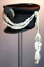 Shako made of black felt, leather top and flap, velvet edge, garnished with white fourragères, safety cords, cockade and metal