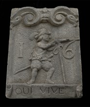 Wall brick from gate with musketeer and ribbon with text qui vive 16, facing stone sculpture sculpture building component