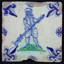 White tile with standing man with gun? in green and blue, corner pattern french lily in blue and edge decoration of rosettes