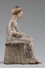 Figurine of a woman. A girl sitting in a chair is dressed in a soft white chiton over which a blue,