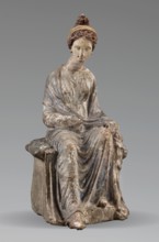 Figurine of a woman. A girl sitting in a chair is dressed in a soft white chiton over which a blue,