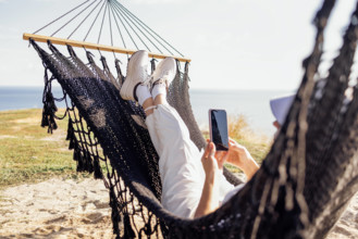 A young woman in casual clothes holds a phone in her hands and lies in a hammock. A girl in a summer hat uses a mobile g